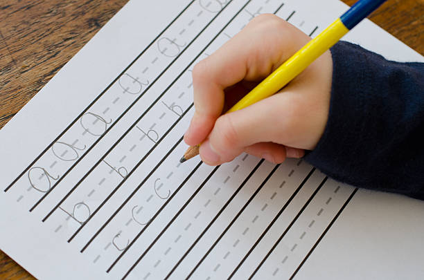 A child's hand holding a yellow pencil, practicing writing on a lined paper worksheet. The paper features dashed lines with letters 'a,' 'b,' and 'c' partially written in a cursive style.