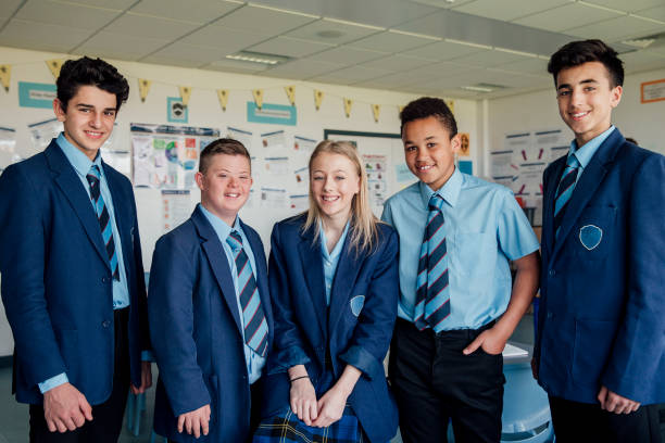 A group of five students wearing school uniforms, consisting of blue blazers, light blue shirts, and ties. They are smiling and standing together in a classroom decorated with educational posters in the background.